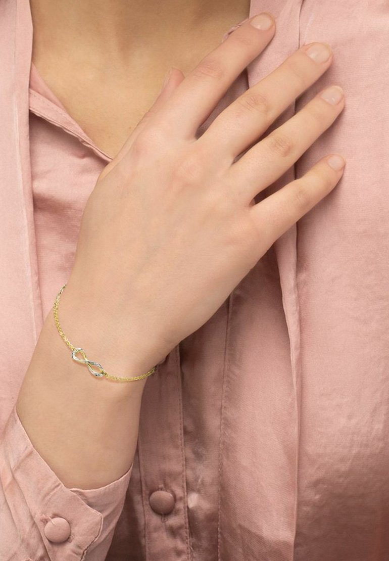 Pulsera de cadena de oro con un acento de símbolo de infinito. La pulsera presenta un diseño delicado, complementado por una blusa de seda de color rosa claro.