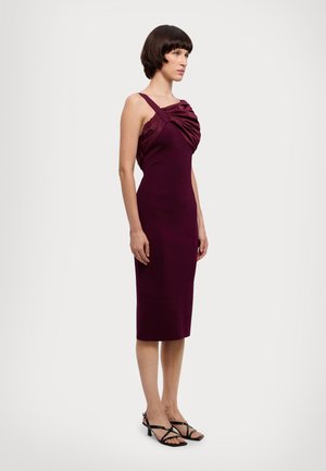 Burgundy fitted dress with a draped satin bust detail, slim silhouette, and adjustable spaghetti straps. Worn with strappy black sandals.
