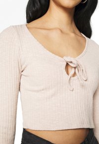 Haut à manches longues côtelé couleur rose clair avec un col en V profond et un accent noué sur la poitrine, doté d'un ourlet court et d'une texture douce.