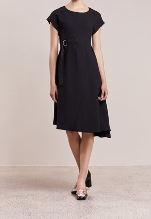 Mujer vistiendo un vestido negro texturizado hasta la rodilla con mangas tipo capa, un dobladillo asimétrico, un cinturón lateral y zapatos negros de tacón grueso con detalles en rosa.