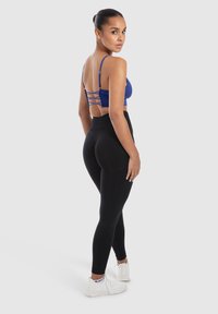 Sujetador deportivo azul con tirantes cruzados en la parte posterior, combinado con leggings negros de cintura alta. La modelo lleva zapatillas deportivas blancas.