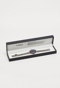 Montre Timex avec un bracelet en maille en acier inoxydable, cadran circulaire bleu avec des accents rouges, marqueurs luminescents et quincaillerie argentée dans une boîte noire.