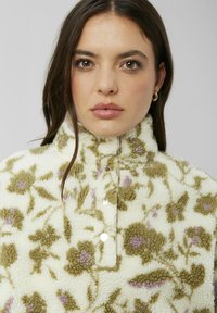Pull en polaire floral crème avec des motifs verts et lavande, col montant et fermeture à boutons-pression. Surface douce et texturée.