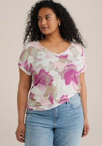 V-neck gėlių marškinėliai su rožine, smėline ir balta abstrakčia_pattern, minkšta medžiaga, trumpomis rankovėmis su apvadu, derinami su mėlynomis džinsinėmis kelnėmis.