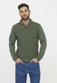 Grüner Pullover aus strukturiertem Material mit Schalkragen, gerippten Manschetten und Saum, kombiniert mit blauen Jeans, steht vor einem neutralen Hintergrund.