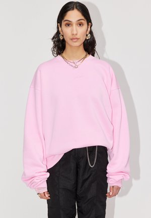 Femme aux longs cheveux foncés portant un sweat-shirt oversize rose clair, un pantalon noir avec une chaîne argentée, des boucles d'oreilles créoles dorées et des colliers superposés.
