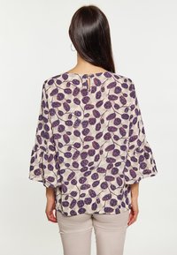 Blusa leggera e trasparente color crema con un motivo a foglie viola, caratterizzata da maniche a campana e un dettaglio a chiusura a oblò sulla schiena.