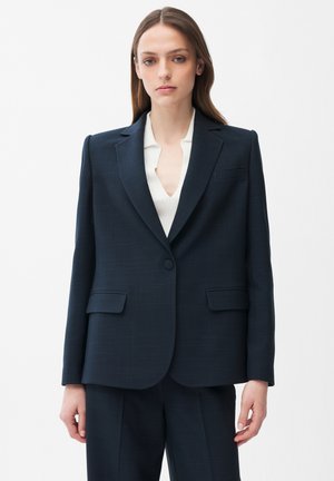 Ung kvinde med glat brunt hår iført en mørkeblå blazer og matchende bukser over en hvid skjorte, stående mod en ensartet hvid baggrund.