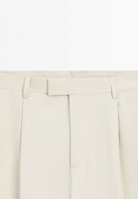 Close-up of beige pants waist with belt loops and a fabric tab covering the front fastening.