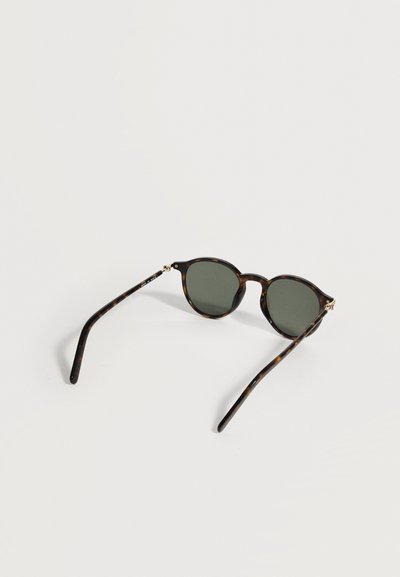 Στρογγυλά γυαλιά ηλίου με ταρταρούγα και σκούρους φακούς που ξεκουράζονται σε λευκή επιφάνεια, εμφανιζόμενα από μια κεκλιμένη πίσω όψη.