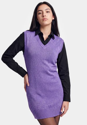 Ung kvinde iført en ærmeløs lilla sweaterkjole over en sort langærmet skjorte, med geometriske øreringe, mod en ensfarvet baggrund.