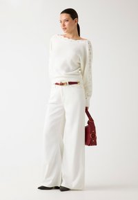 Pull en tricot blanc avec taille côtelée, pantalon blanc à jambes larges, ceinture bourgogne et sac à main, avec des accents en métal doré.