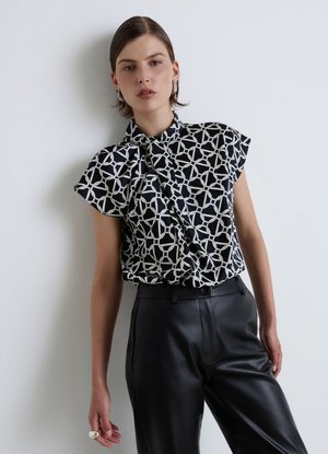 Donna che indossa una camicetta con motivo geometrico bianco e nero a maniche corte e pantaloni in pelle neri a vita alta, in piedi contro un muro bianco.