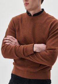 Suéter de cuello redondo marrón con textura, con puños y dobladillo de canalé, usado sobre una camisa de color oscuro con cuello. La tela presenta un patrón tejido.