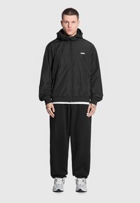 Młody mężczyzna stoi na jednolitym tle, ubrany w czarną bluzę na zamek, czarne dresy i trampki, z widocznymi tatuażami na rękach.