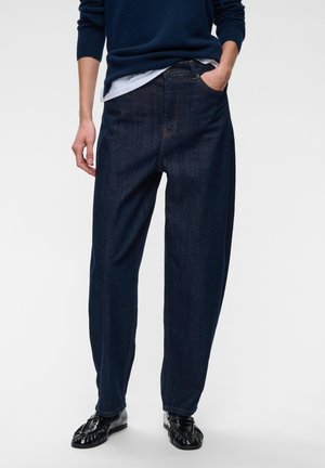 Personne portant un jean bleu foncé ample avec une main dans la poche, associé à des chaussures noires et un pull bleu foncé.