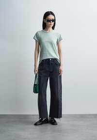 Γυναίκα με μαύρα γυαλιά ηλίου, ριγέ πράσινο T-shirt, φαρδιά σκούρα τζιν με φθαρμένα τελειώματα, μαύρα loafers, κρατώντας μικρή πράσινη τσάντα, όρθια σε γκρίζο πάτωμα.