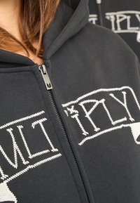 Sweat à capuche noir zippé en coton, doté d'un texte brodé blanc et d'un logo sur le devant. Comprend un tissu texturé et une fermeture éclair en métal.