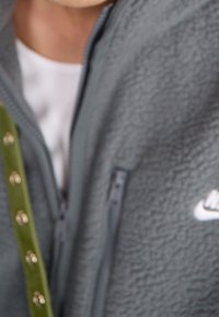 Grå fleecetröja med hög krage, dragkedjefickor och en vit Nike-logotyp. Grön rem med gulddetaljer fäst.