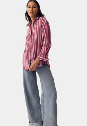 Kvinde iført en løs rød- og hvidstribet skjorte med opkrogede ærmer og lyseblå jeans med vide ben mod en ensfarvet baggrund.