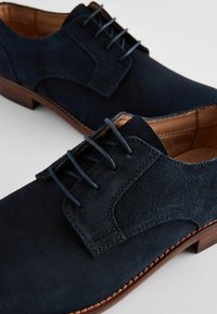 Zapatos de ante azul marino con cordones, de textura suave, con costuras en contraste y suelas de cuero marrón. Presentan un diseño clásico y acentos mínimos.