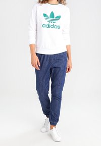 Sweat à texture blanche avec le logo Adidas vert et trois bandes, associé à un pantalon navy ample et des baskets blanches.