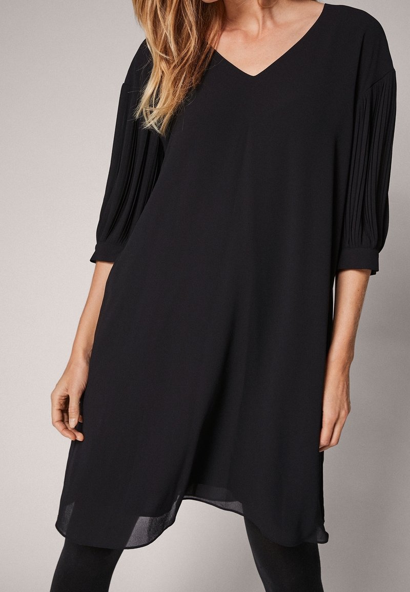 Femme portant une robe noire ample arrivant aux genoux avec un col en V et des manches trois-quarts plissées sur un fond neutre.