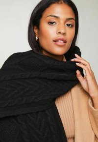 Sort kabelstrik scarf draperet over en beige ribbet top, der viser teksturerede mønstre. Modellen holder tørklædet foran en neutral baggrund.