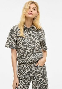 Korte ermer skjorte og bukser sett i beige med svarte zebra-striper. Har krage og brystlomme, laget av et glatt stoff.