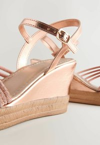 Sandalias de cuña en oro rosa con un acabado metálico suave, que presentan correas entrelazadas y una suela tejida texturizada para añadir agarre.