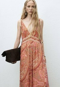 Largo vestido fluido en un patrón de paisley rosa y dorado, con un escote en V profundo y acentos de encaje. Sostiene un bolso clutch de ante marrón oscuro.