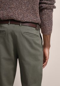 Olive green trousers with two back pockets, paired with a brown leather belt. The model wears a textured brown sweater.