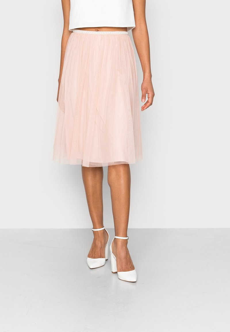 Femme portant une jupe midi en tulle rose clair et des talons blocs blancs à bride à la cheville, debout devant un fond blanc uni.