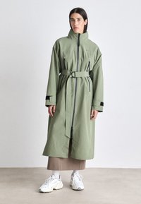 Cappotto lungo, con cintura, in tessuto verde oliva chiaro. Presenta un colletto alto, una zip frontale e due tasche con zip. Sneakers bianche visibili sotto.