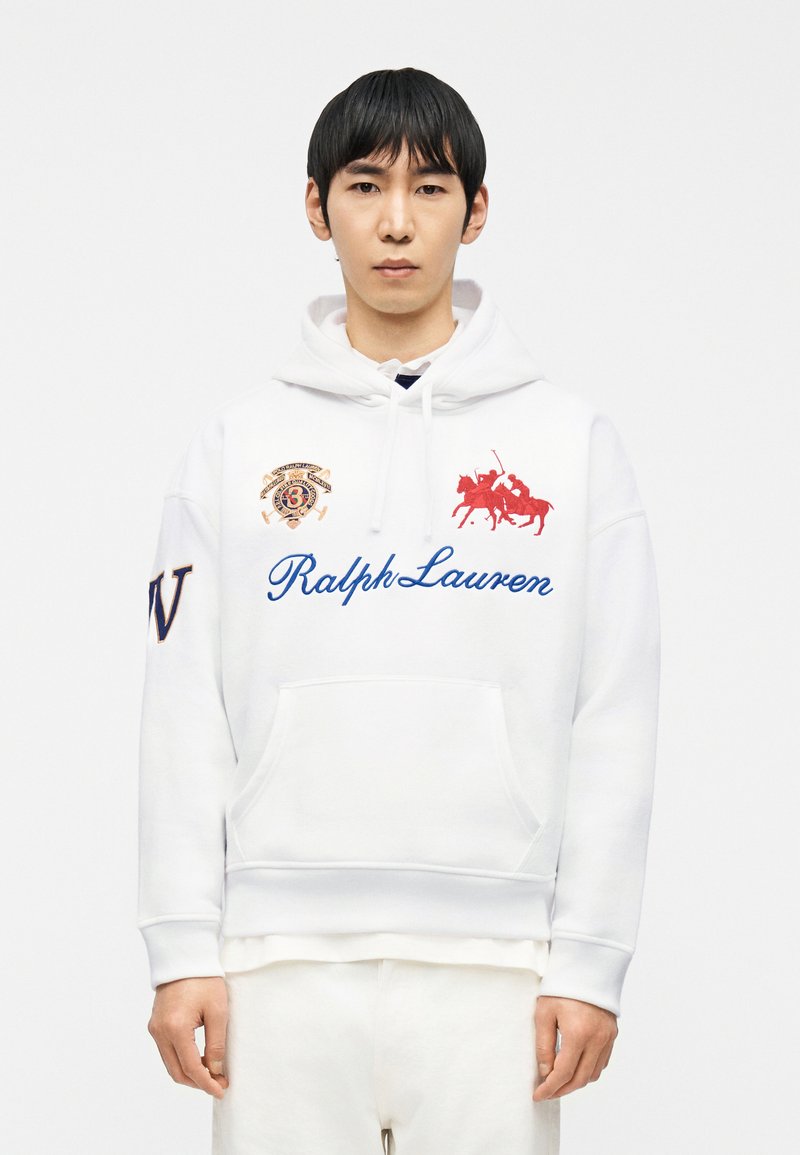 Νεαρός άνδρας που φοράει λευκό φούτερ Ralph Lauren με  μπλε γράμματα, κόκκινο λογότυπο παίκτη πόλο και κεντημένα μπαλώματα στο στήθος και το μανίκι.