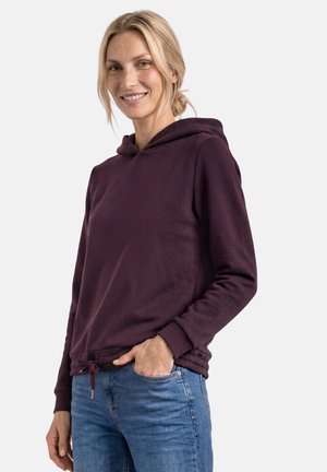 Frau mit blondem Haar in einem niedrigen Dutt, die einen dunkelvioletten Hoodie und blaue Jeans trägt, lächelt mit einer Hand in der Tasche.