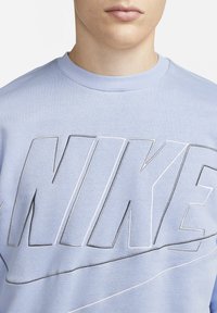 Světlomodrý svetr s velkým vyšitým logem "NIKE" v bílé a černé barvě, s klasickým kulatým výstřihem a měkkou texturou.