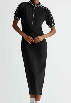 Femme portant une robe midi noire à manches courtes, avec une fermeture éclair à l'avant, un col et des poignets de marque, ainsi qu'un liseré blanc sur les épaules.