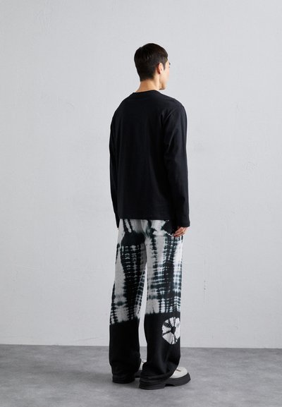 Schwarzes Langarmshirt kombiniert mit weit geschnittenen, schwarz-weißen Batik-Hosen, die ein markantes Muster und einen lässigen Schnitt aufweisen. Klobige Schuhe.