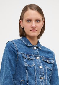 Denimjakke med blå finish, klassisk krage, to brystlommer og sølvfargede knapper for lukking. Jevn tekstur og enkel design.