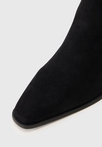 Stivale alla caviglia in suede nero con punta appuntita e suola bassa, mostrato su uno sfondo bianco.