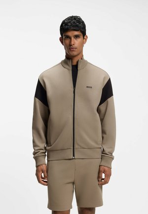 Uomo che indossa una giacca beige con zip e pannelli neri sulle spalle e pantaloncini beige abbinati, in piedi davanti a uno sfondo bianco semplice.