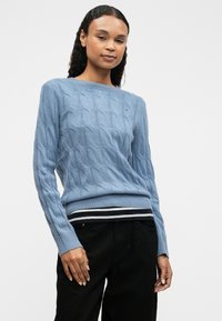 Svetlo modrý sveter s pleteným vzorom, okrúhlym výstrihom, rebrovanými manžetami a pruhovaným lemom. Vhodne skombinovaný s tmavými nohavicami pre kontrast.