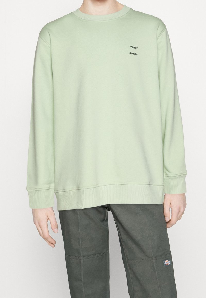 Sweatshirt vert clair avec encolure ronde, poignets et ourlet côtelés. Présente deux petits logos noirs imprimés sur la poitrine.