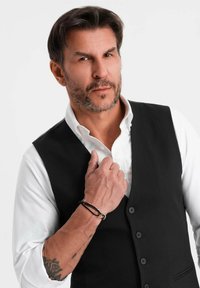 Uomo con capelli corti scuri e barba, vestito con una camicia bianca e un gilet nero, che tiene il risvolto del gilet mostrando un tatuaggio e braccialetti sul braccio sinistro.