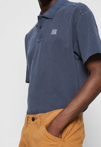 Dunkelblaue Baumwoll-Poloshirt mit kragenförmigem Ausschnitt und kurzen Ärmeln, versehen mit einem kleinen Logo-Patch. Kombiniert mit beigen Hosen.