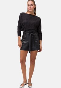 Schwarze Ledershorts mit hoher Taille und Bindegürtel-Detail, kombiniert mit einem dunklen, funkelnden Langarmoberteil und silbernen Absätzen.