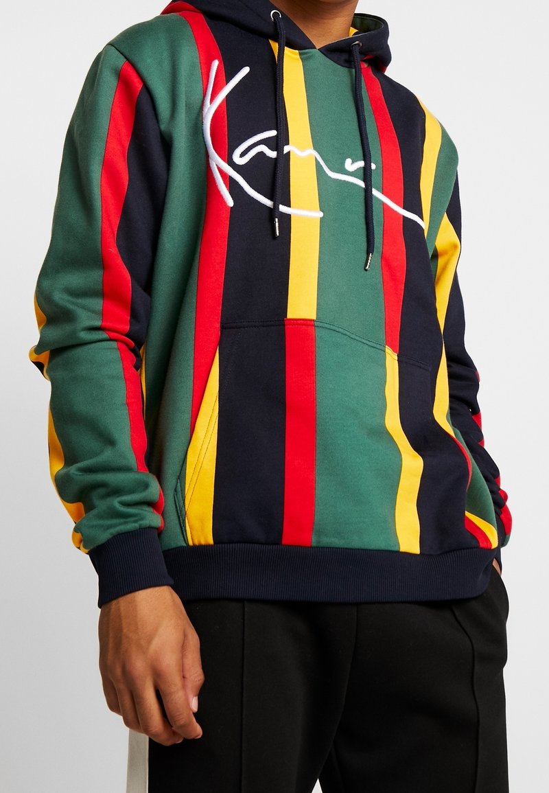Hombre vistiendo una sudadera con capucha con rayas verticales verdes, rojas, amarillas y negras, y un texto en blanco en el pecho, combinado con pantalones negros.