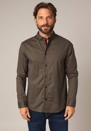Chemise marron à boutons avec un motif à pois subtil, manches longues, et boutons noirs. Portée avec un jean bleu, sur fond neutre.