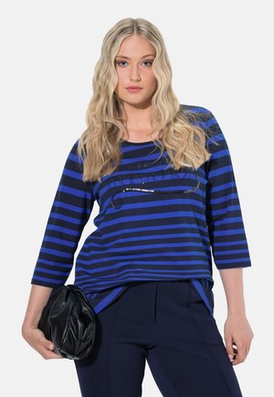 Blauw en marineblauw gestreepte top met lange mouwen en een ronde halslijn, met pailletten versiering op de borst. Model houdt een zwarte tas vast.